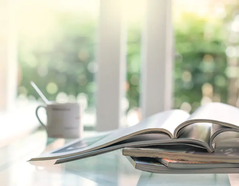 Aufgeschlagenes Magazin liegt auf einem Glastisch neben einer weißen Kaffeetasse mit Löffel, im Hintergrund helle Fenster mit grünem Ausblick.